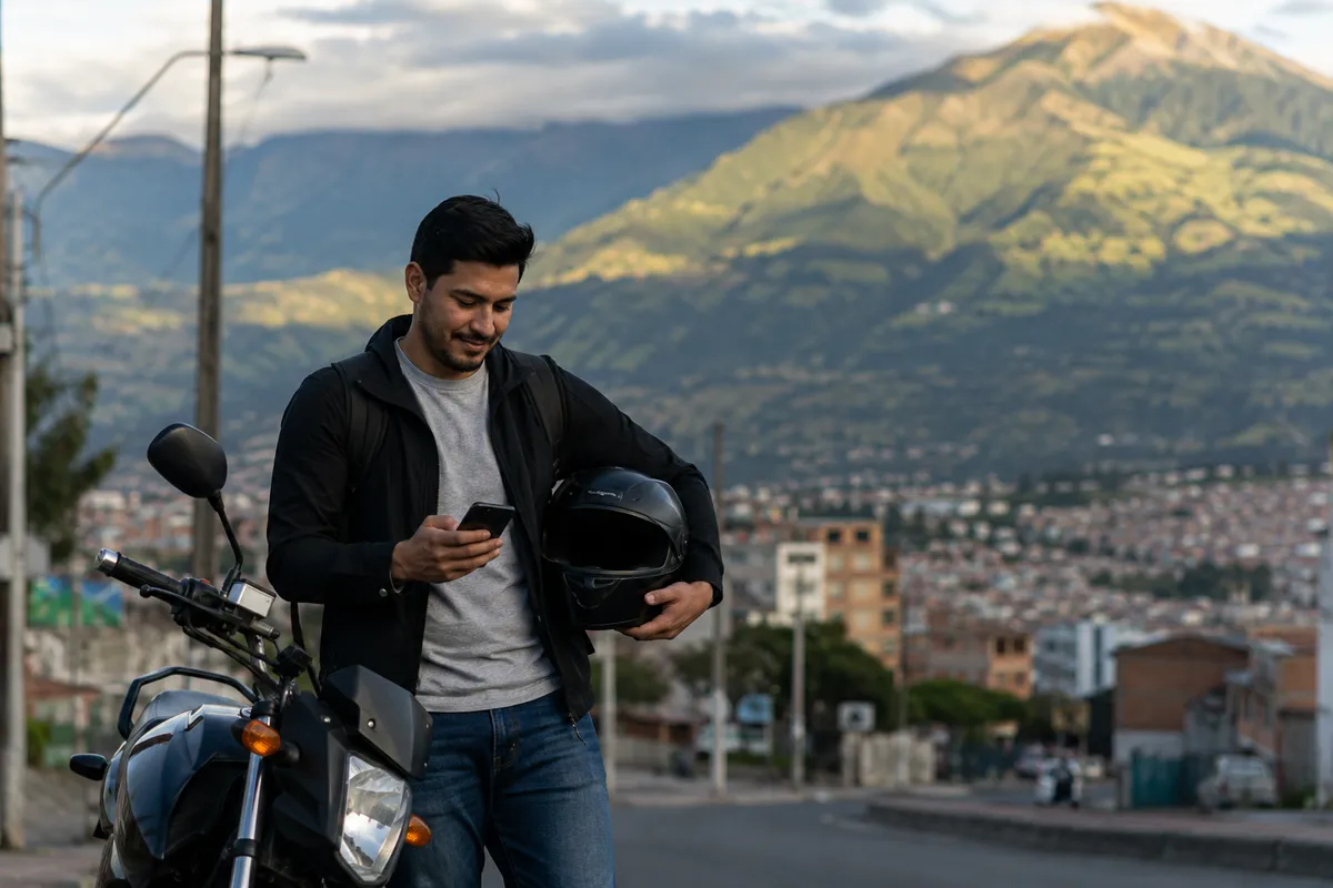Motociclista revisando su celular junto a su moto, con Pasto y el volcán Galeras al fondo